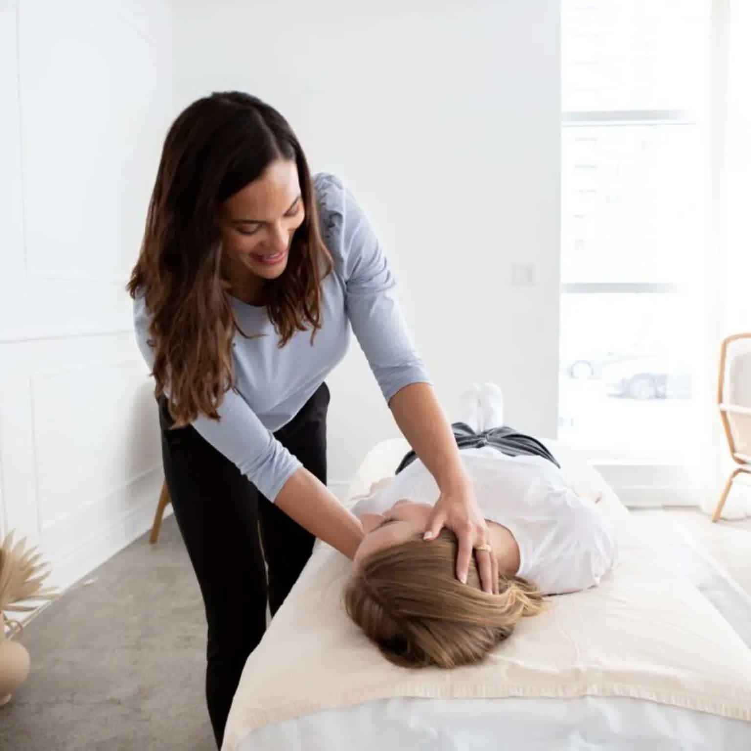 Woman assisting another person on a bed, hands on their shoulders in a bright, calm room.