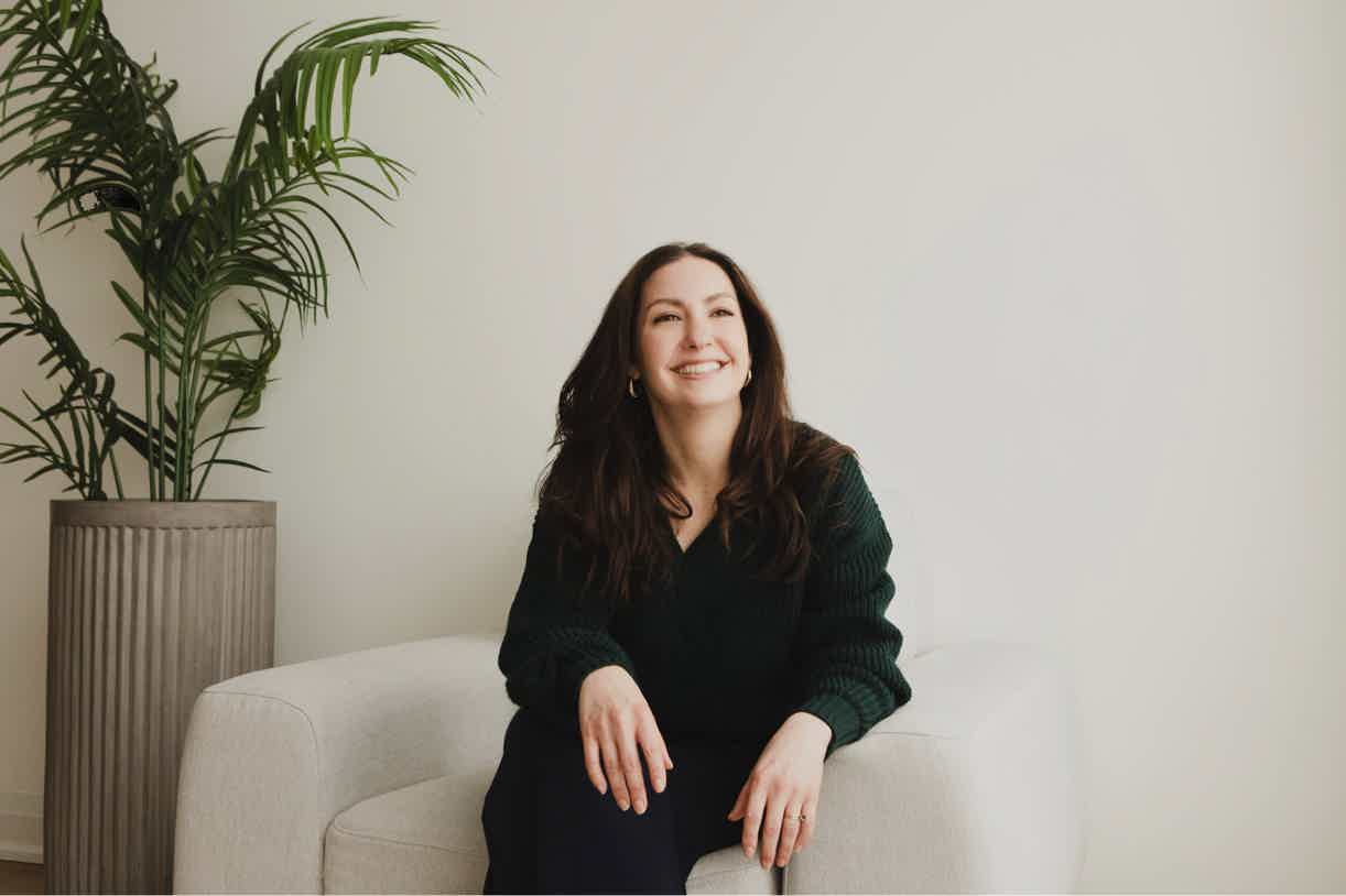 Smiling woman in dark outfit seated on a light sofa next to a potted plant against a white wall