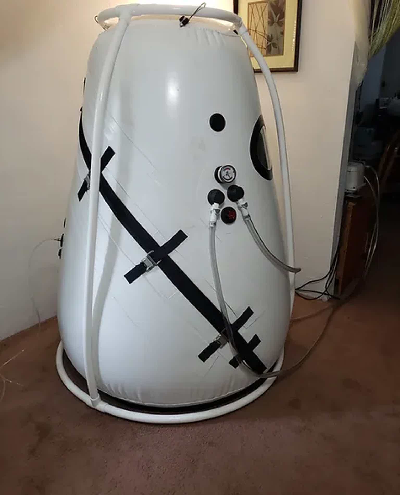 White, egg-shaped hyperbaric chamber with control valves and straps, standing in a room.