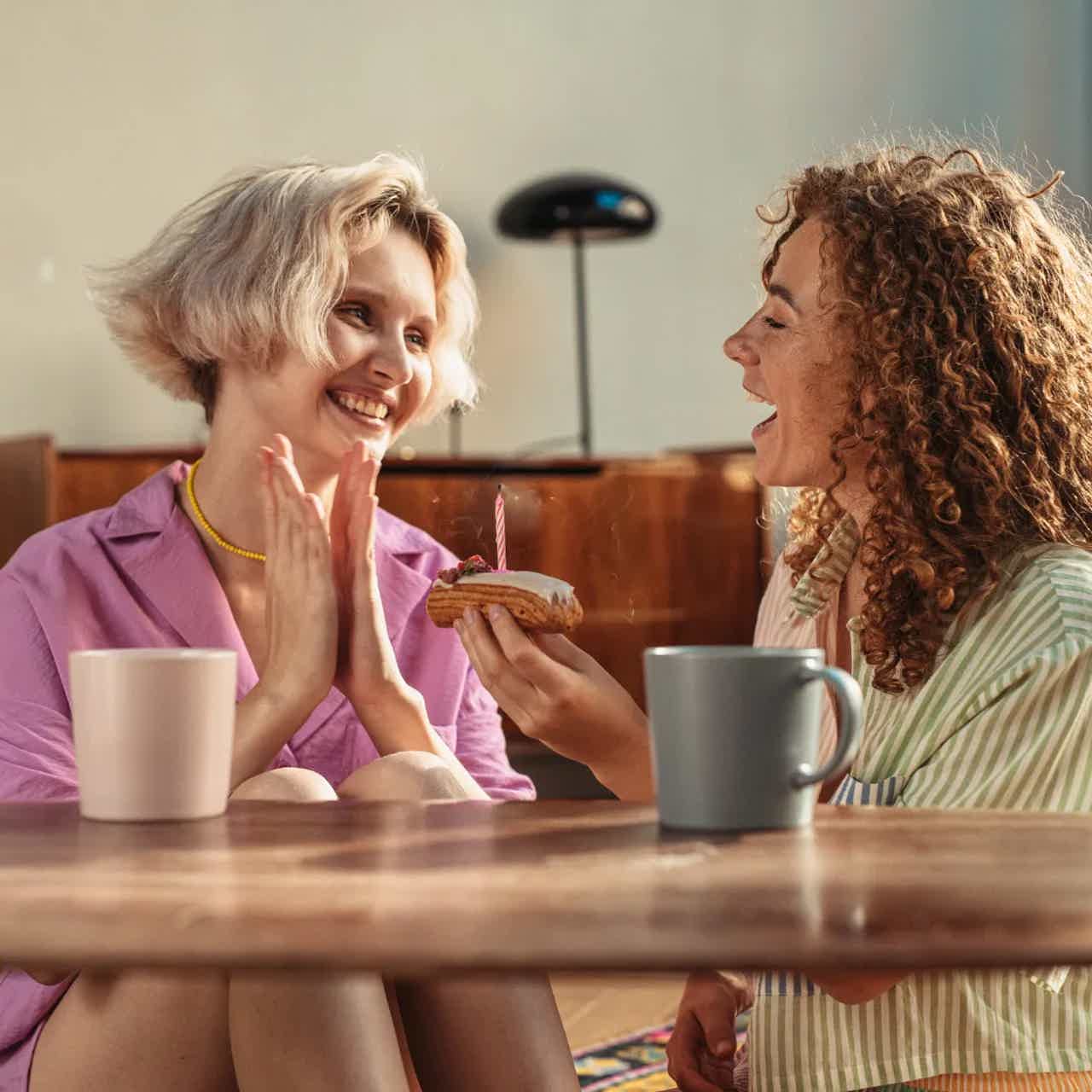 Two women celebrate a birthday at a cafe; one holds a cupcake with a candle while the other claps and smiles.