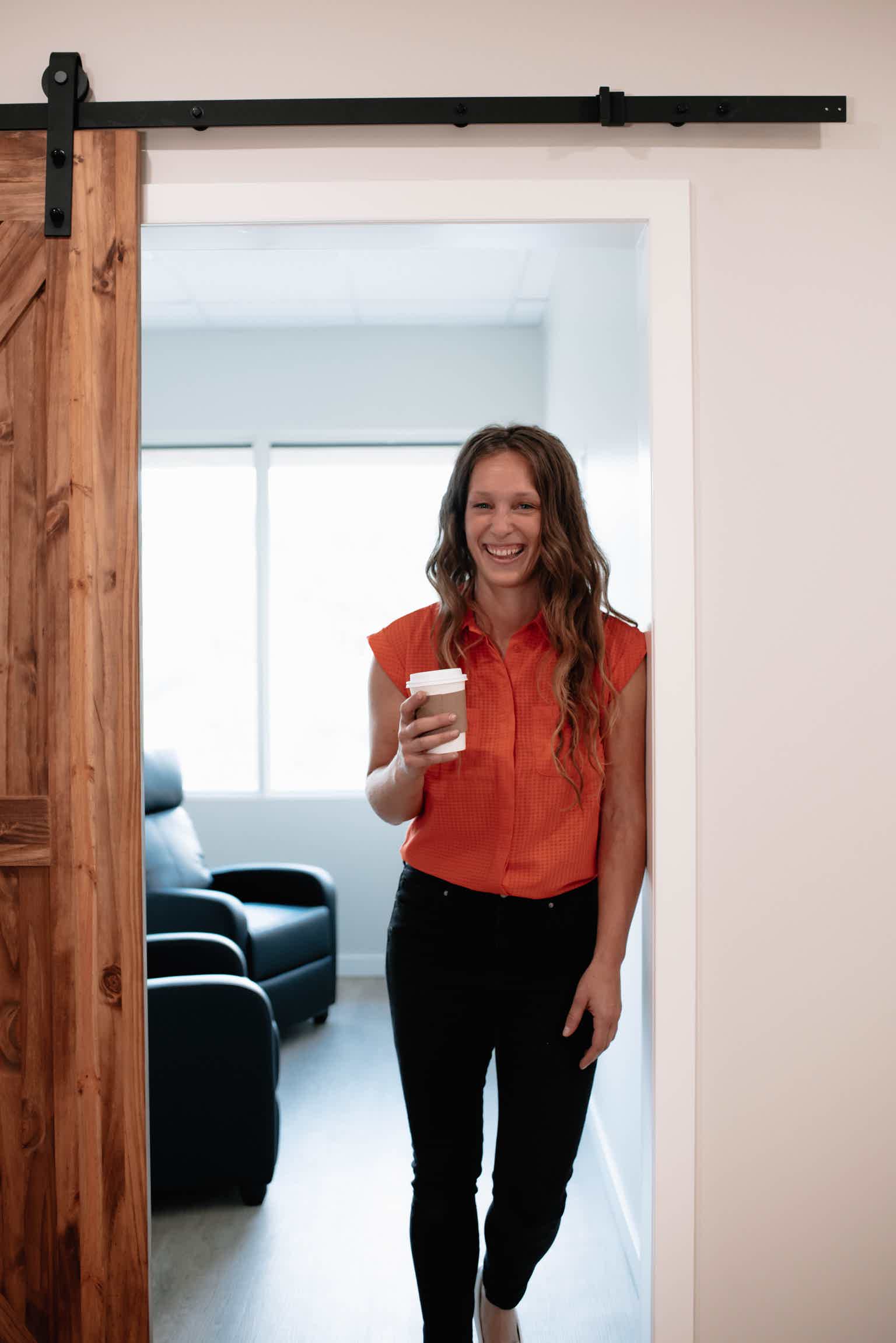Woman in orange shirt holding coffee, standing in doorway of bright living room. Sliding barn door on track.