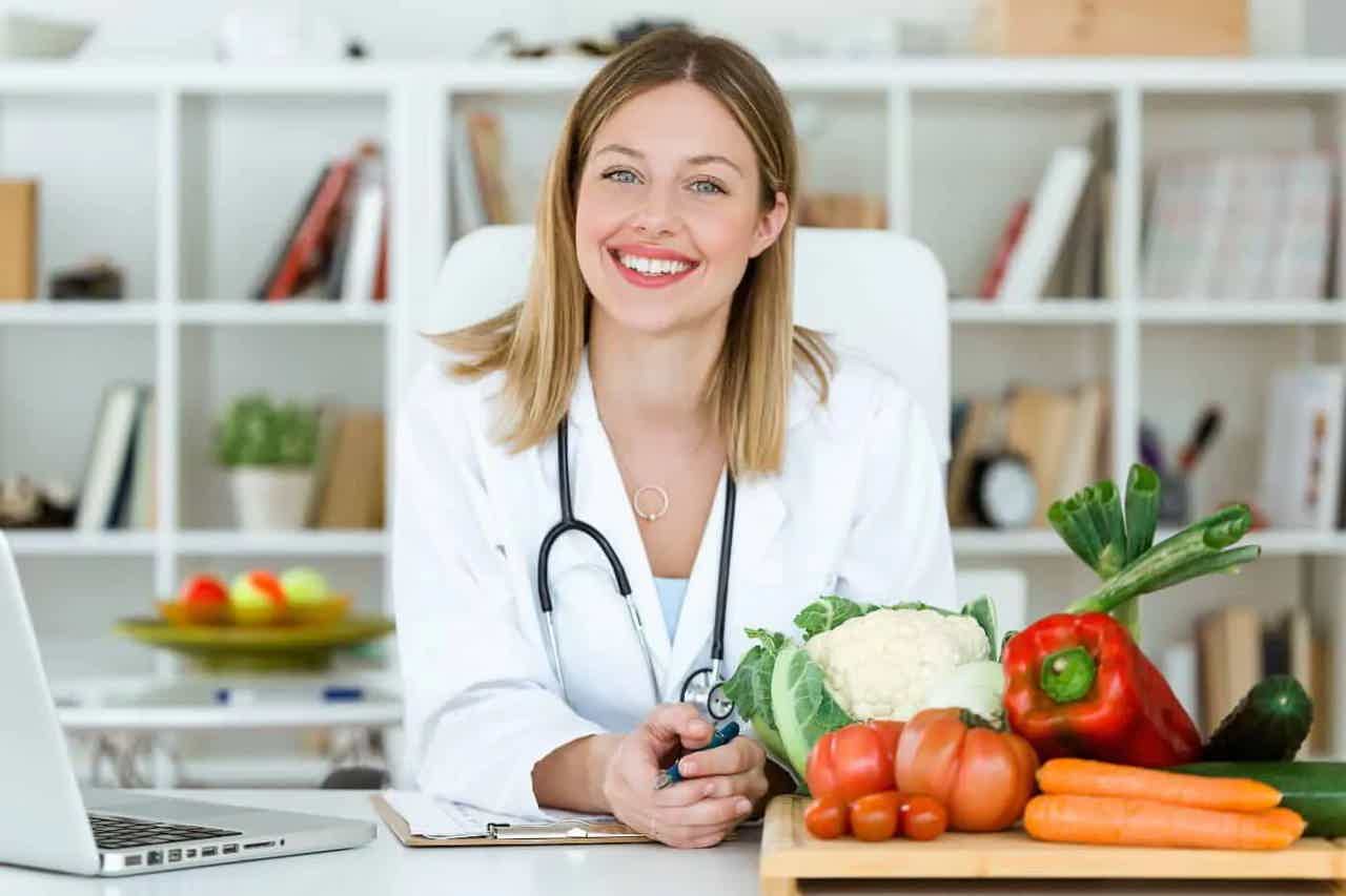 Smiling doctor in white coat holding stethoscope, beside assorted fresh vegetables on a countertop.