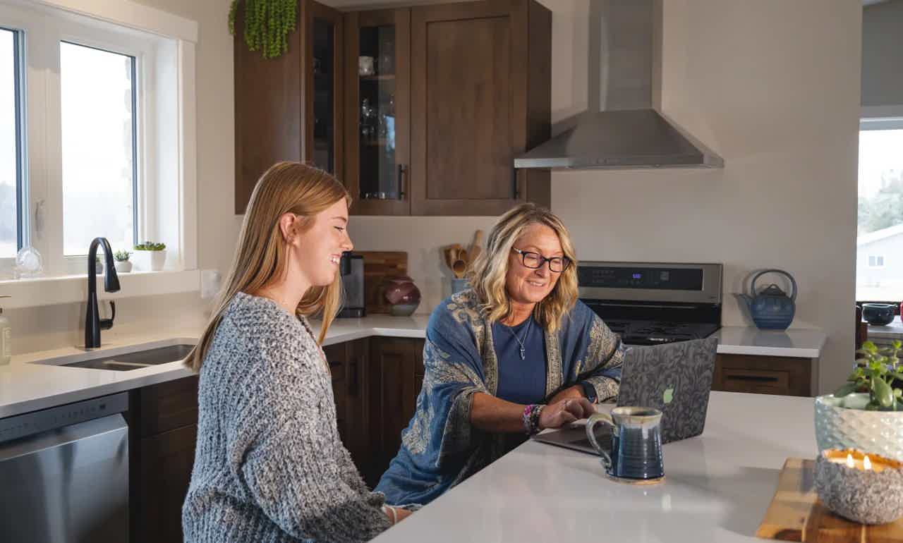Two women chat at a kitchen island, one pouring tea, in a bright modern home.