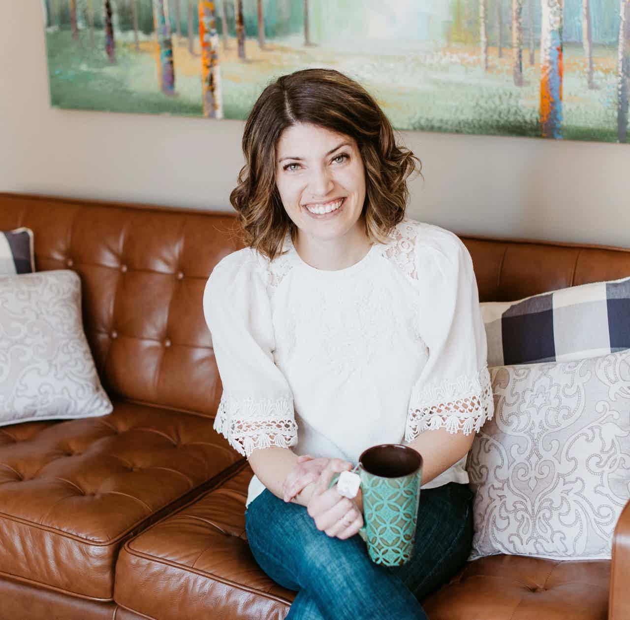 Smiling woman with wavy brown hair sits on brown leather couch, holding a teal mug, in a cozy living room.