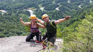 Two climbers with helmets pose joyfully on a rocky ledge, forested canyon below.