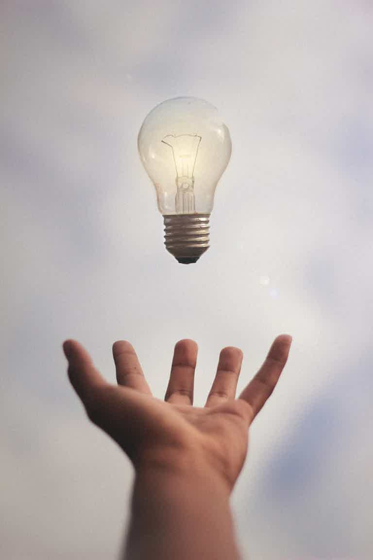 Hand beneath a glowing light bulb hovering above, sky in soft focus.