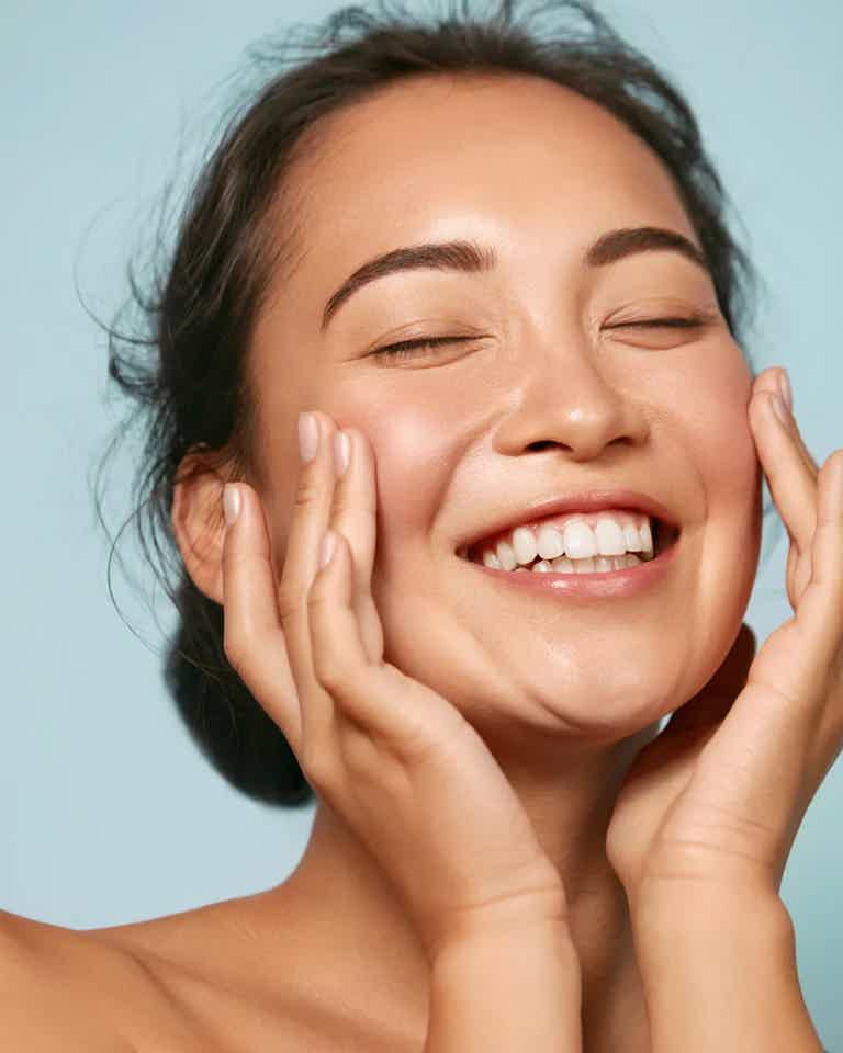 Smiling woman with hands gently cupping cheeks, eyes closed, showcasing clear skin against light blue background.