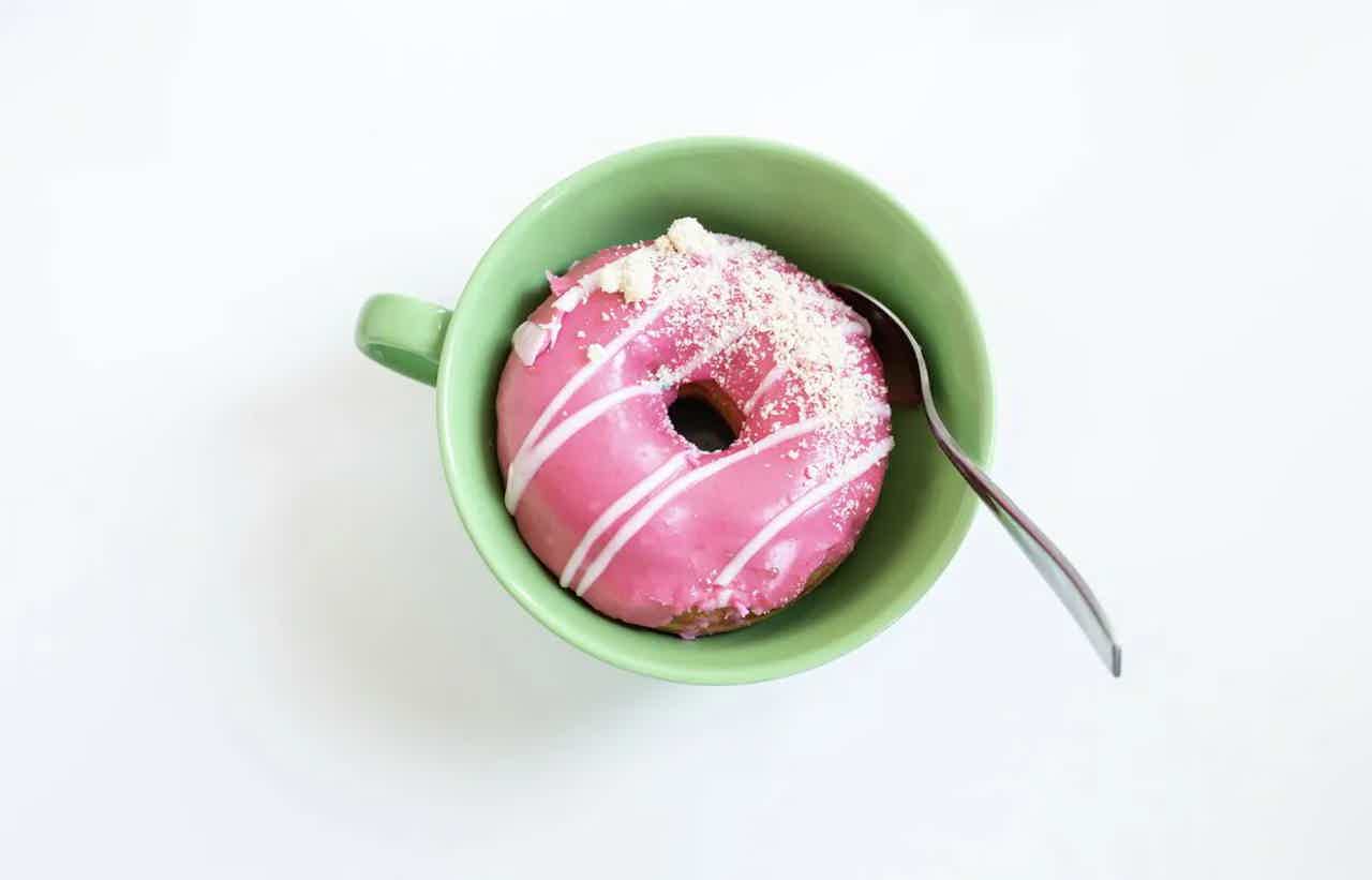 Pink frosted donut with sprinkles in a green bowl, with a spoon beside it on a white background.