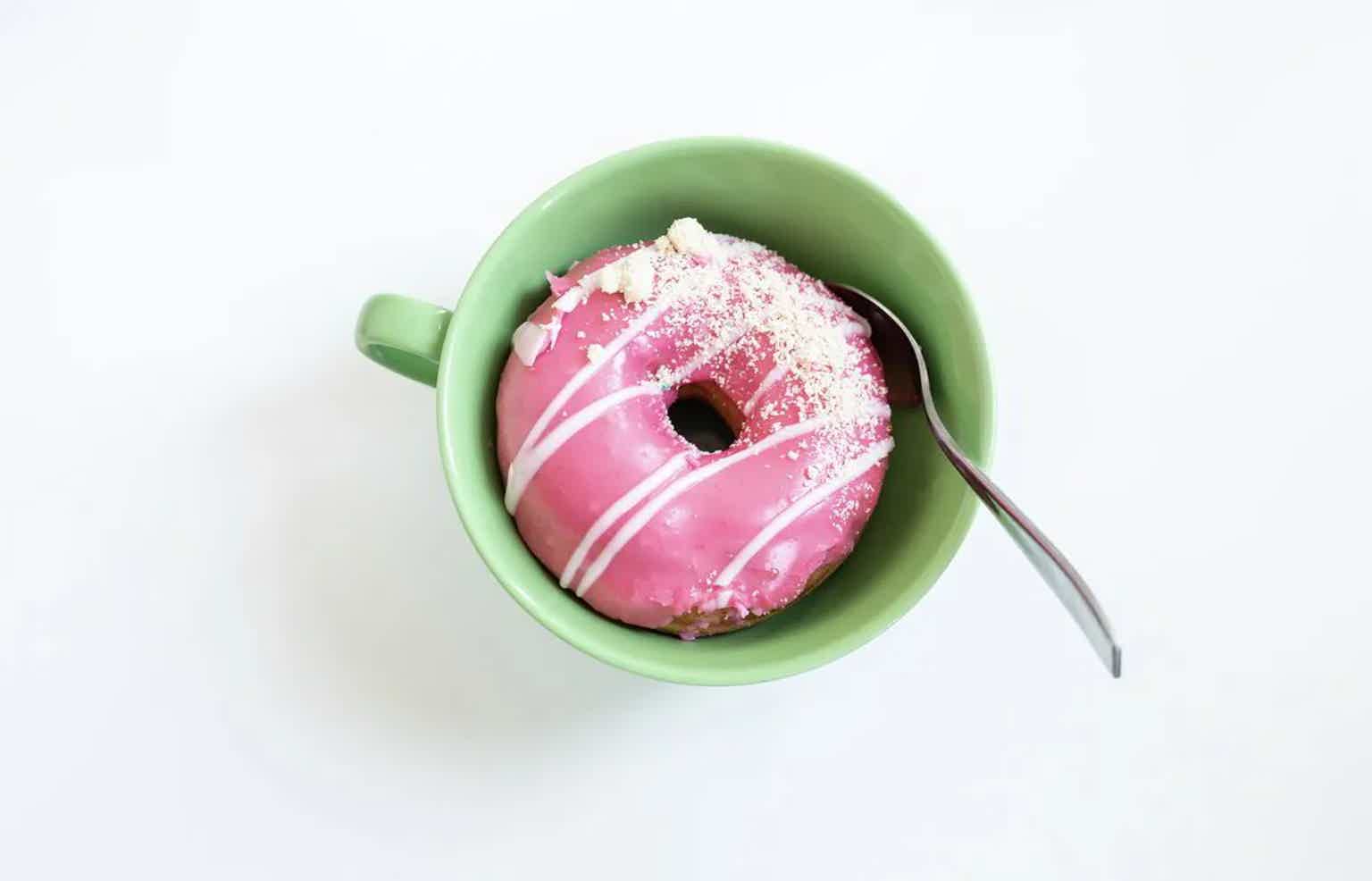 Pink frosted donut with sprinkles in a green bowl, with a spoon beside it on a white background.