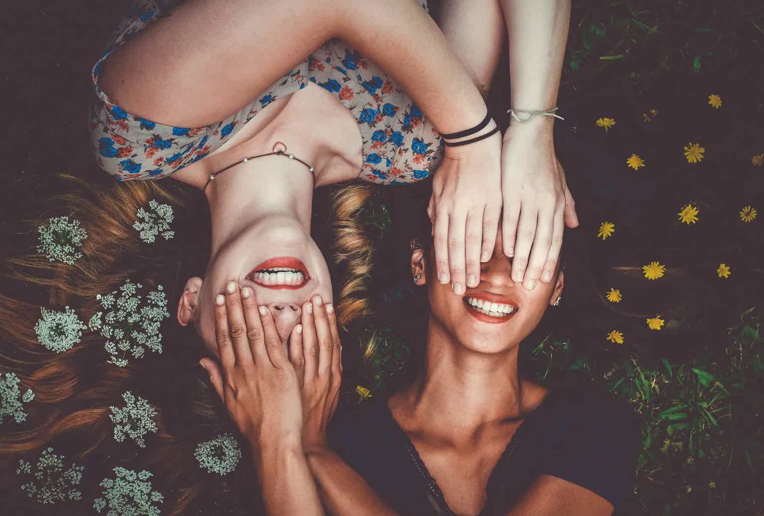 Two women lie on grass, smiling with hands covering their faces playfully amid flowers.