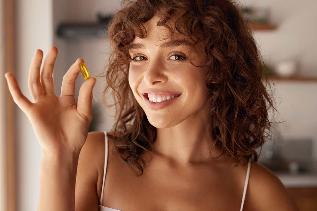 Curly-haired woman smiling and waving with gold ring in cozy indoor kitchen setting.