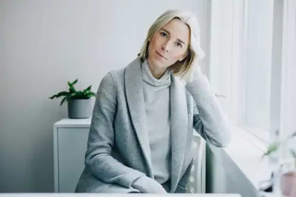 Blonde woman in gray blazer and turtleneck seated by a bright window, resting head on hand and looking toward camera