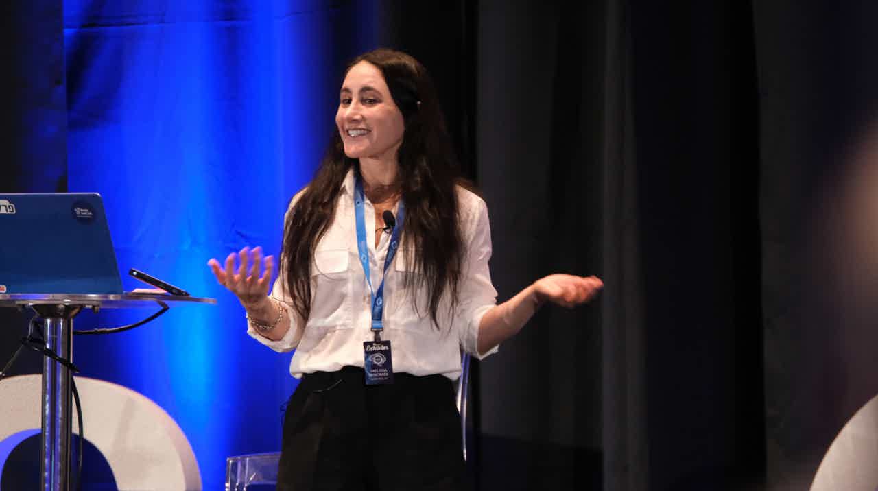 Speaker on stage gesturing with hands, wearing white blouse and lanyard, standing near laptop on podium.