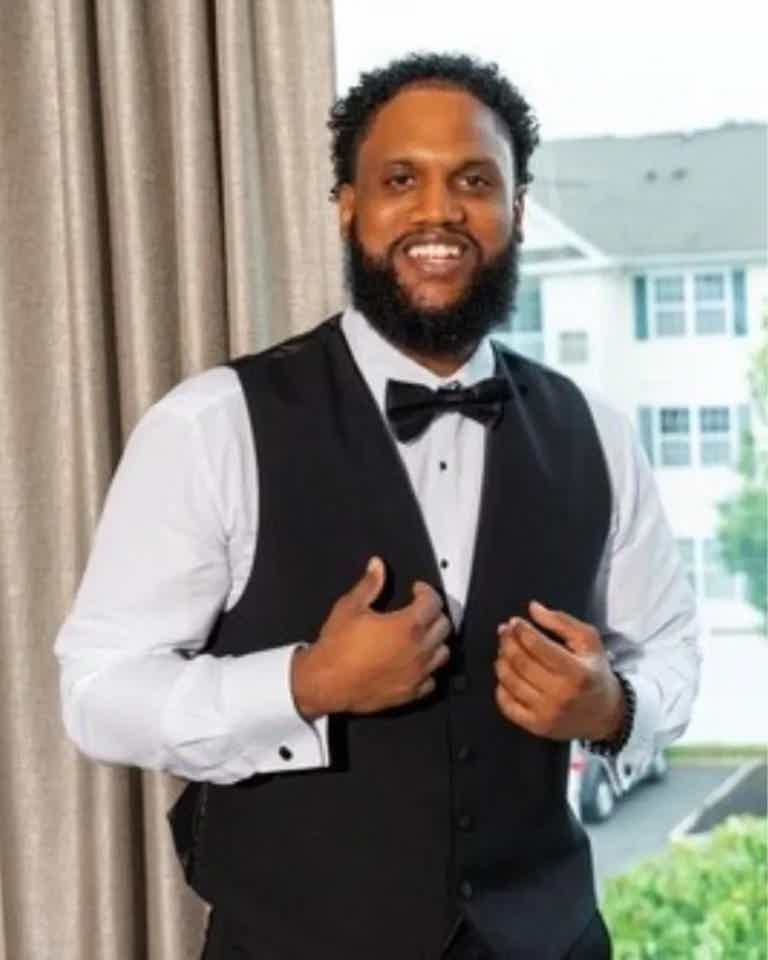 Man in white dress shirt, black vest and bow tie, smiling, standing indoors near curtains.