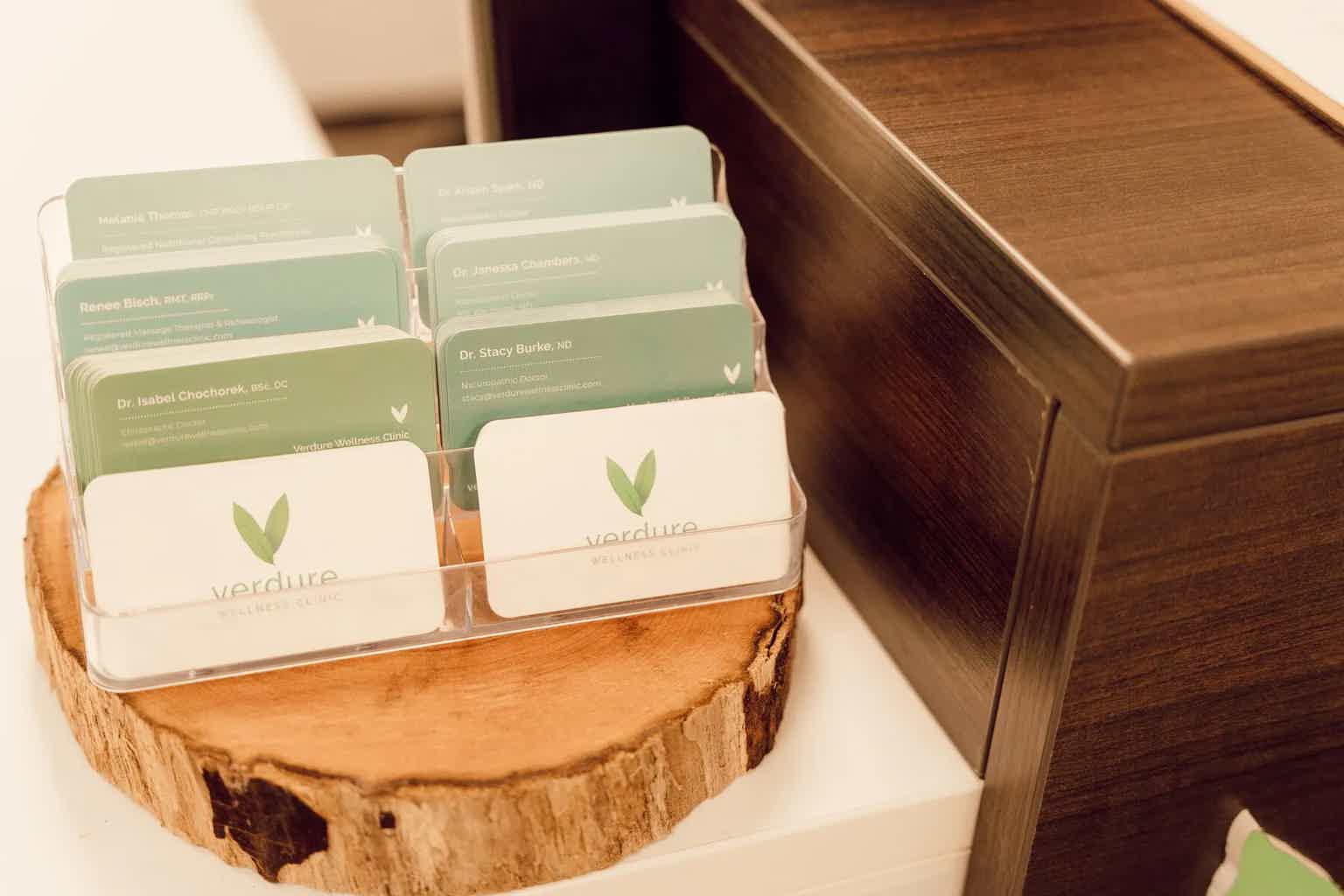 Verdure Wellness Clinic name cards in a clear acrylic holder on a wooden countertop, with leaf logos and doctor names.