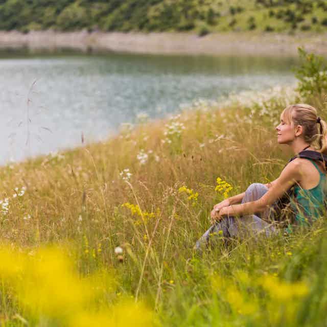 Woman sits in grassy meadow by a lake, gazing peacefully into the distance.