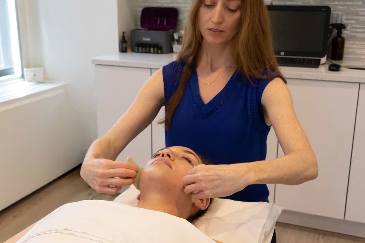 Practitioner in blue treats a client’s face on a treatment table using gua sha stones.
