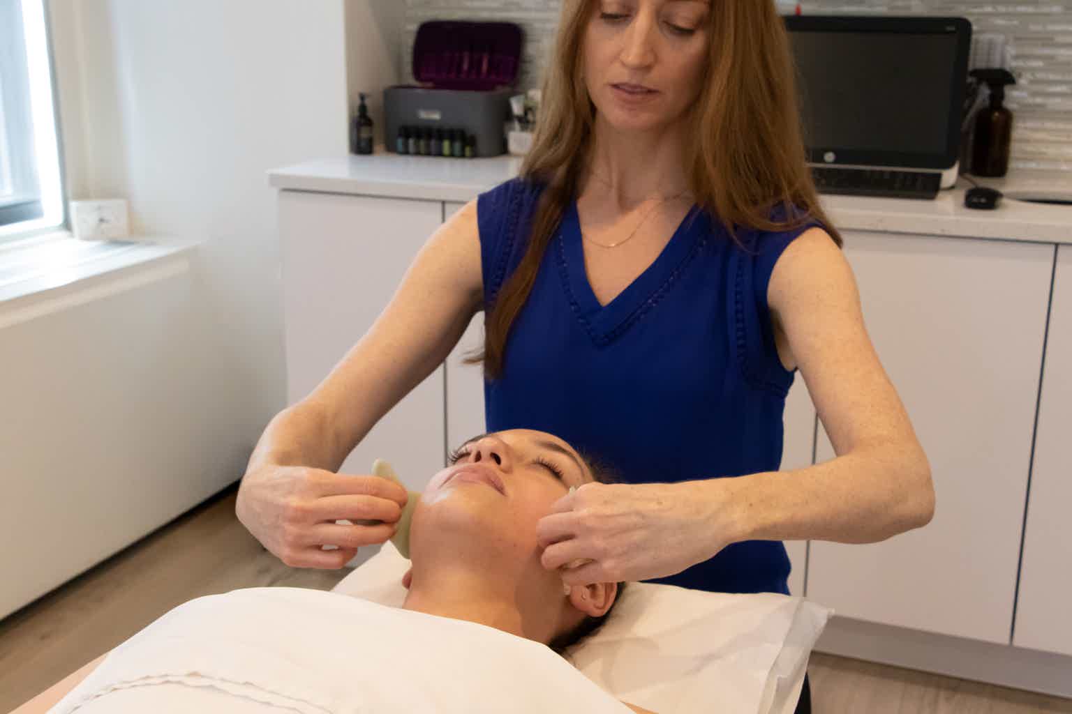 Practitioner in massages client’s face on a treatment table using gua sha stones.