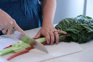 Person chopping celery stalks on a cutting board; leafy greens nearby, knife slicing celery.