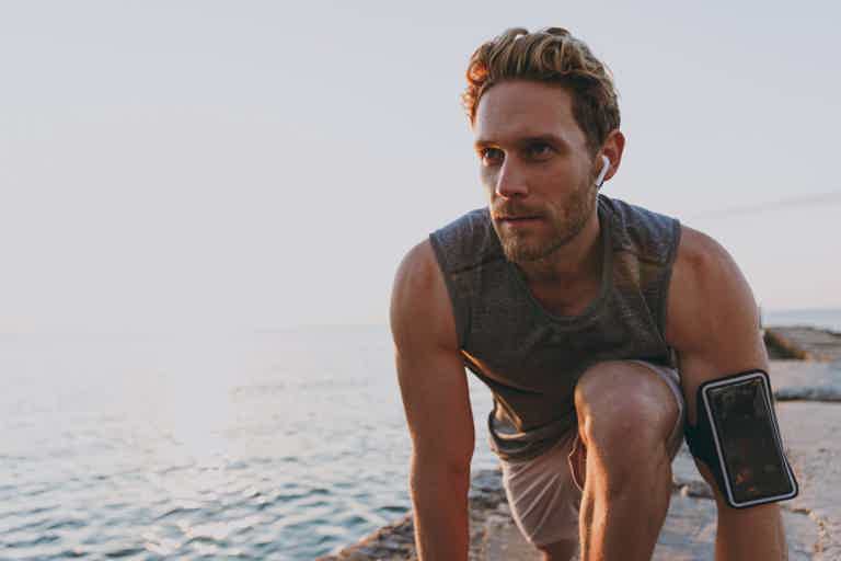 Athletic man jogging by the ocean, wearing a fitness band/phone strapped to his arm, crouched forward at dusk.
