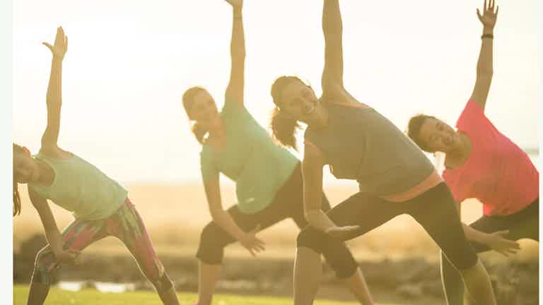 Group of people doing outdoor stretching/yoga with arms raised in a sunny setting.