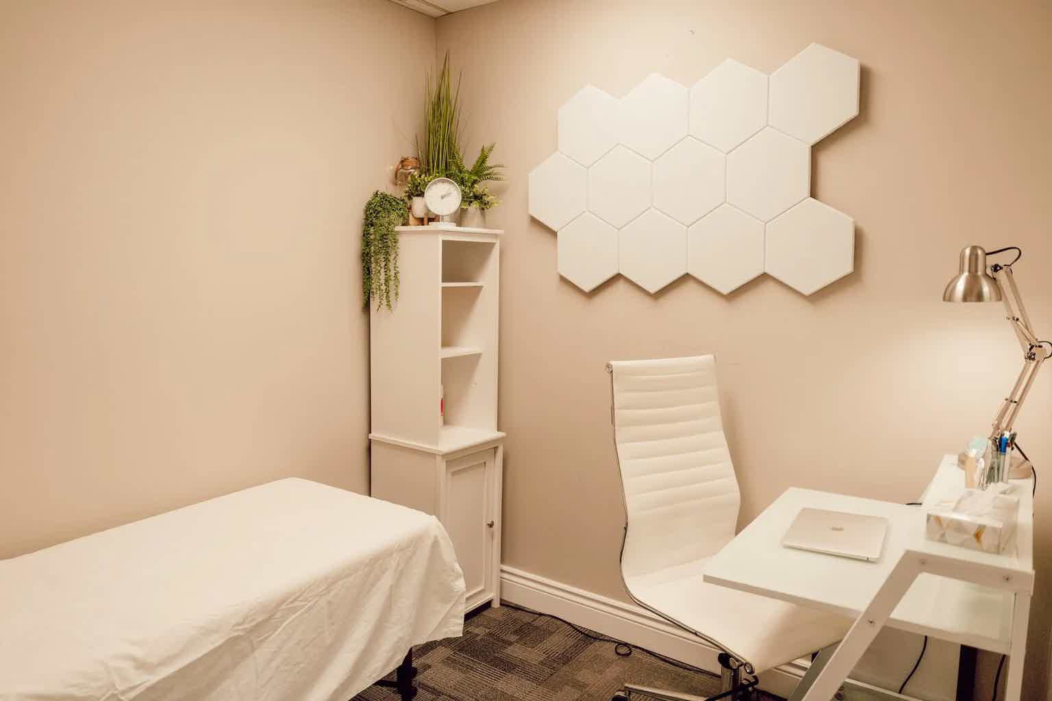 Clean treatment room with white massage table, shelving unit, mounted hexagon wall decor, and plant arrangement on wall.