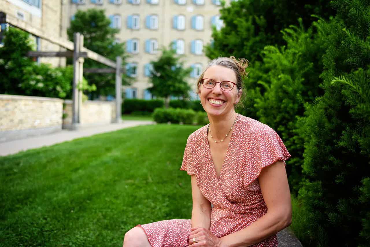 Smiling woman with glasses in a pink dress sits on a path beside green bushes and a stone building.