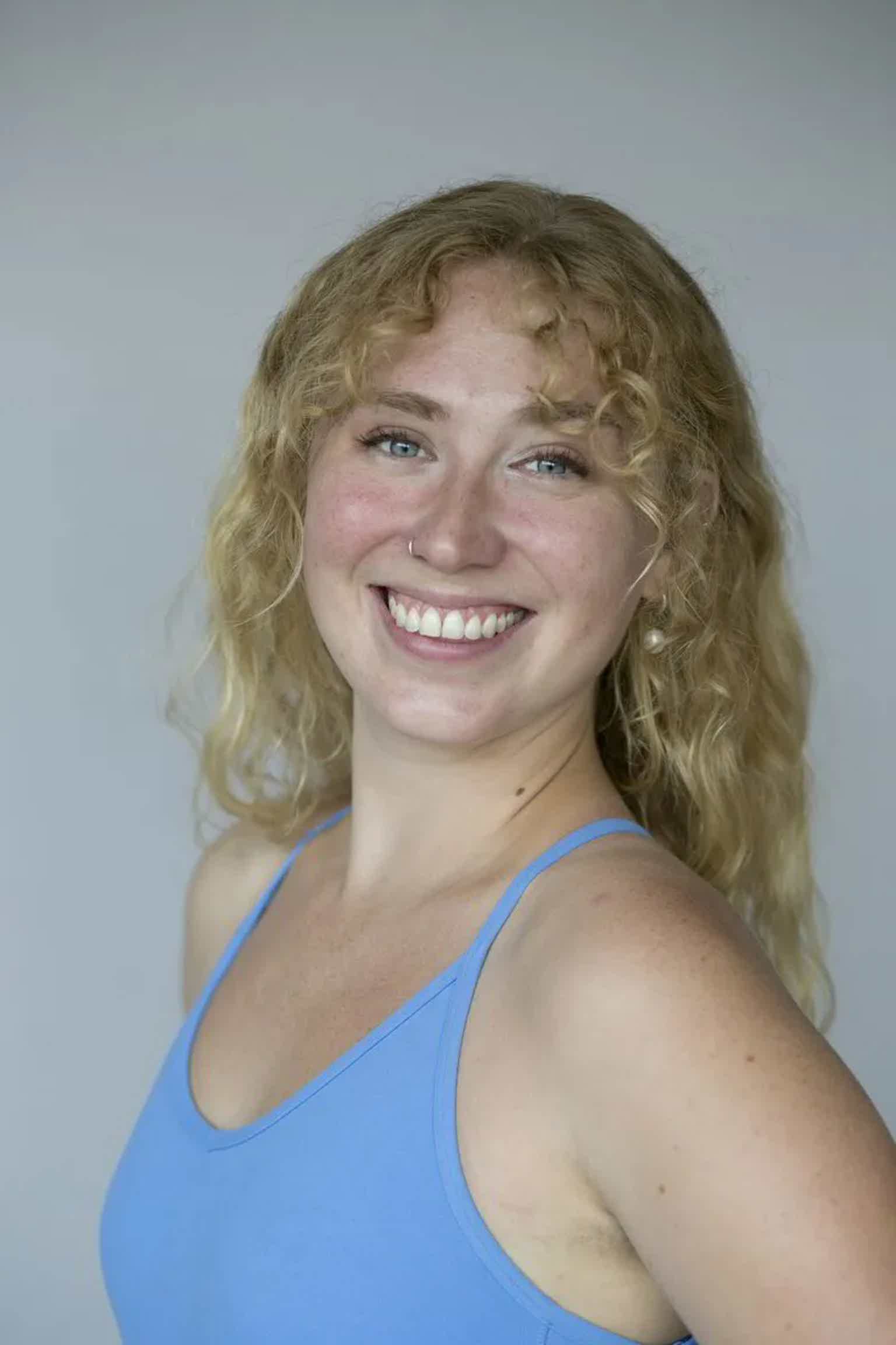 Smiling blonde woman with curly hair wearing a blue tank top, facing camera against a plain light background.