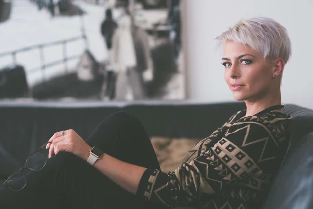 Woman with short platinum hair sits on a couch, gazing thoughtfully, wearing a patterned sweater.