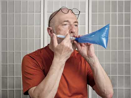 Older man wearing glasses and red shirt blowing into a small blue mouthpiece in a tiled room