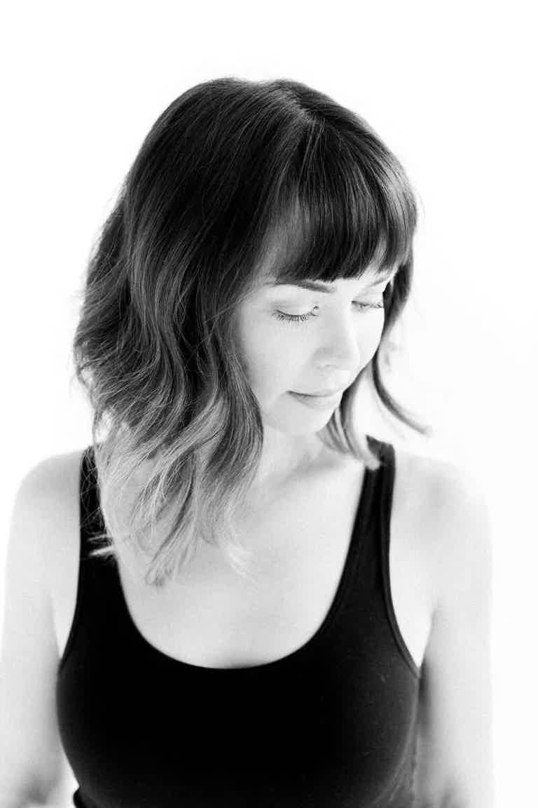 A woman with wavy hair and bangs looks downward, captured in a soft black-and-white portrait against a light background.