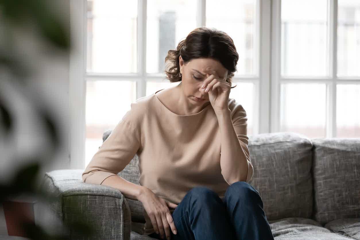 Woman sitting alone on a sofa, head down and hand pressed to her face in visible distress, suggesting mood symptoms of estrogen imbalance.