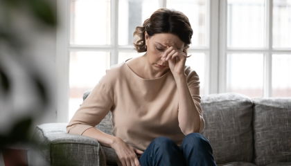 Woman sitting alone on a sofa, head down and hand pressed to her face in visible distress, suggesting mood symptoms of estrogen imbalance.