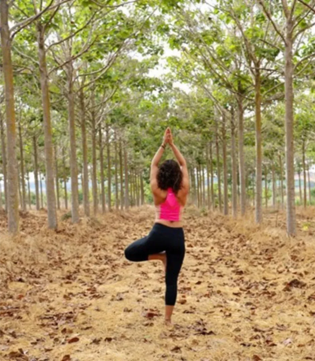 Woman in pink sports bra and black leggings doing tree pose in a tree-lined path with fallen leaves.