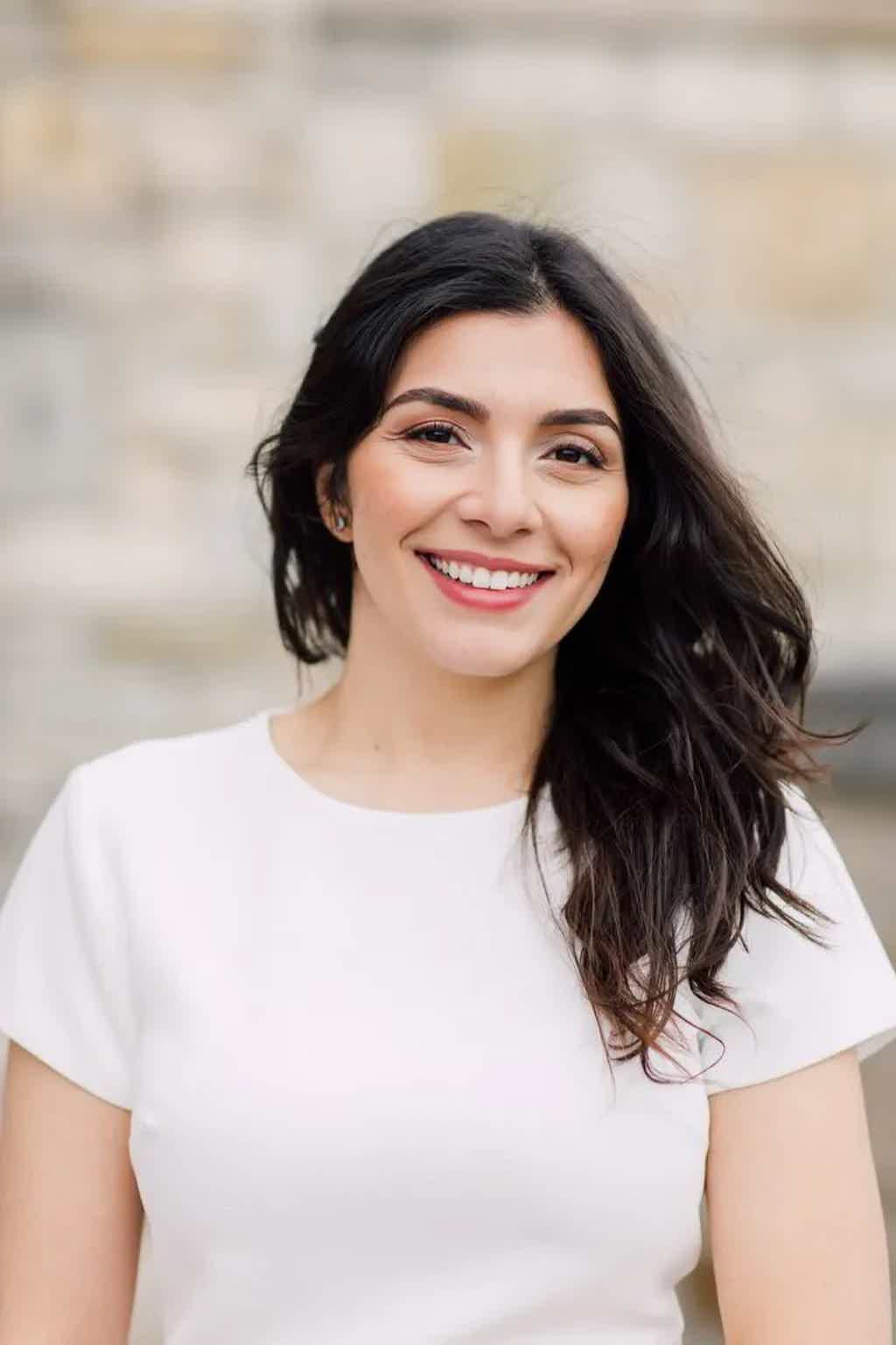 Smiling woman with dark wavy hair wearing a white top, posed outdoors against a blurred background.