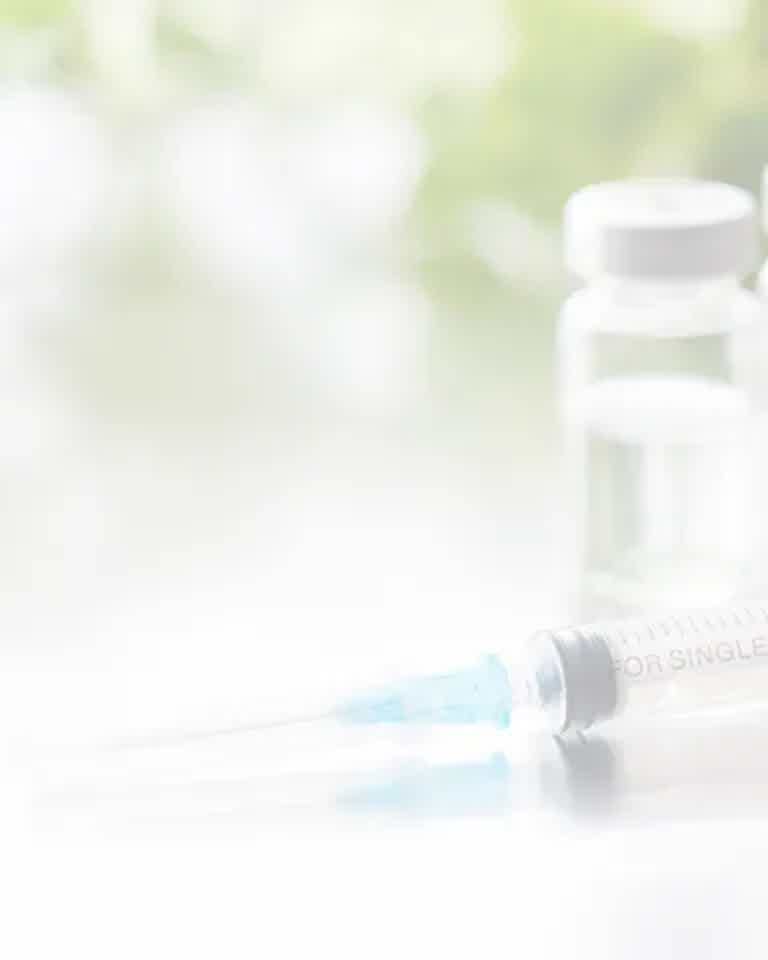 Syringe with blue needle cap and small medical vial on a brightened surface in soft-focus background.