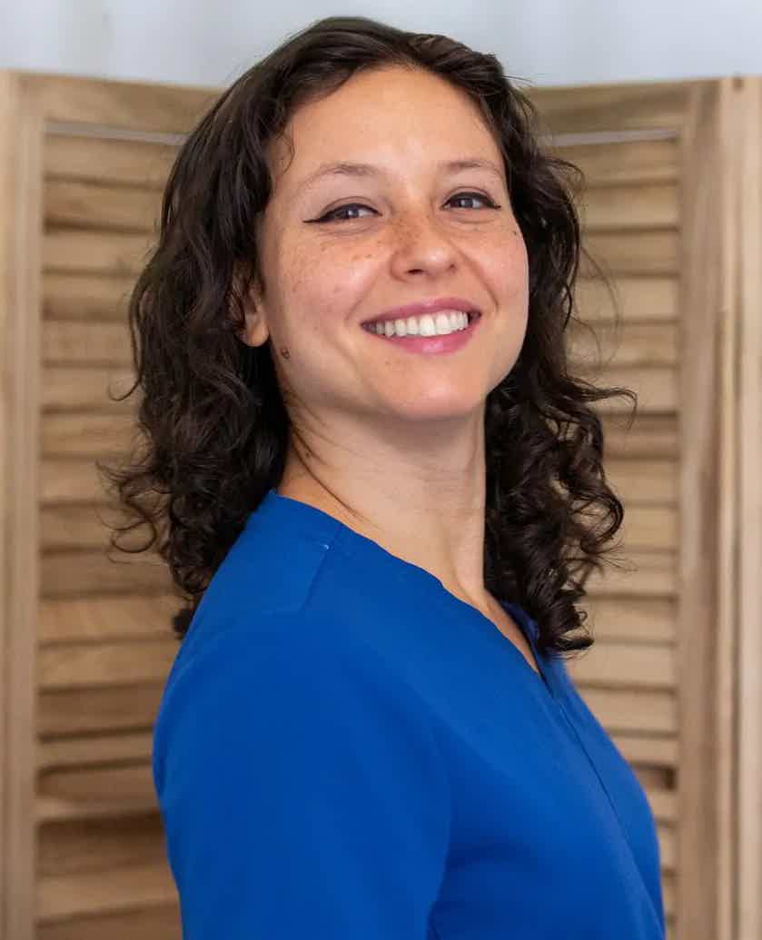 Smiling woman in blue blouse standing sideways indoors with wooden room divider behind her.