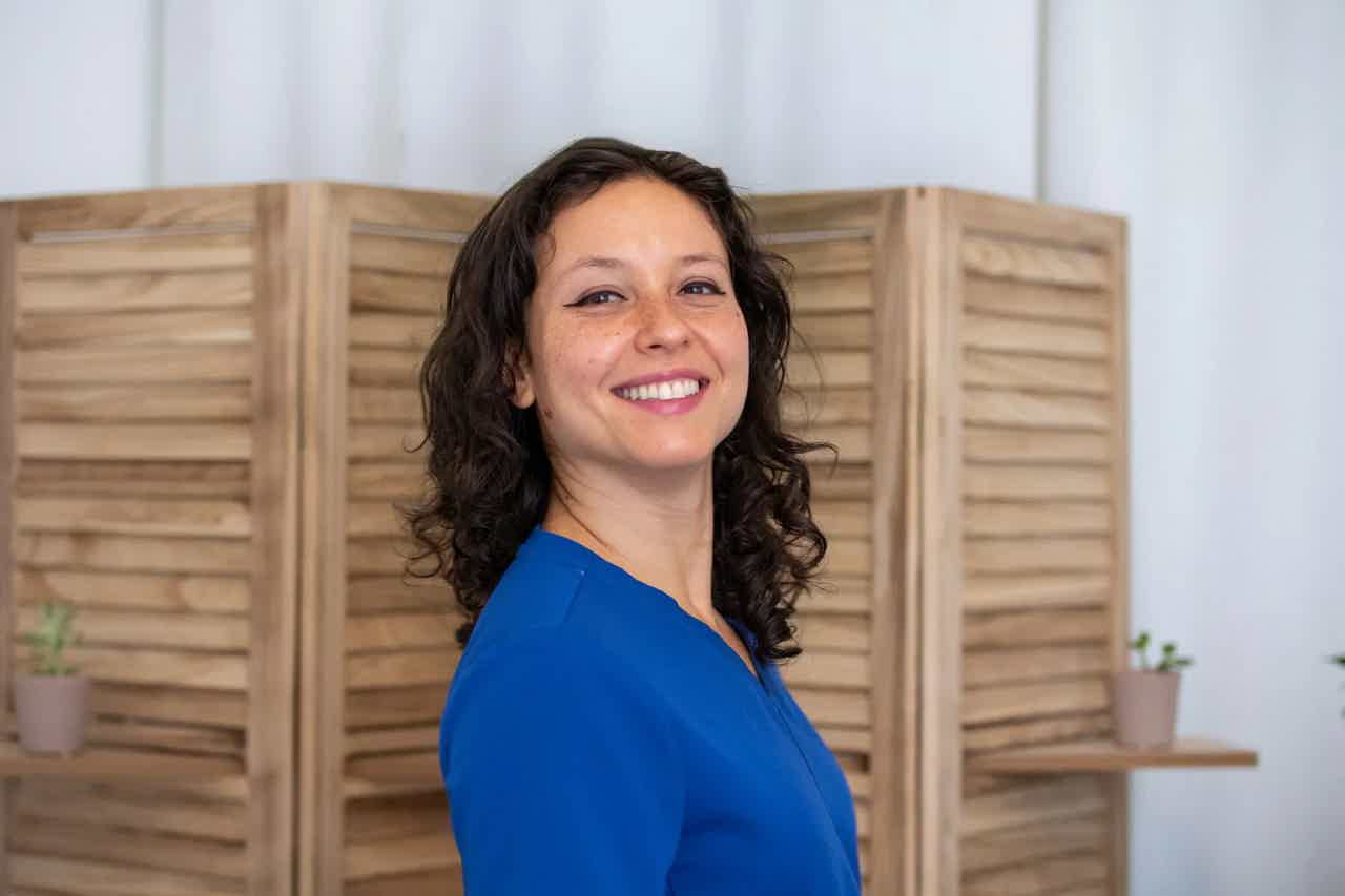 Smiling woman in blue blouse standing sideways indoors with wooden room divider behind her.