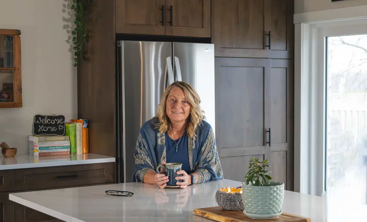 Smiling woman seated at kitchen island holding a mug, with cabinets, plants, and window in background.