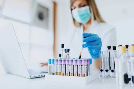 Masked lab technician in blue gloves pipetting a hormone sample into a rack of test tubes during clinical analysis.