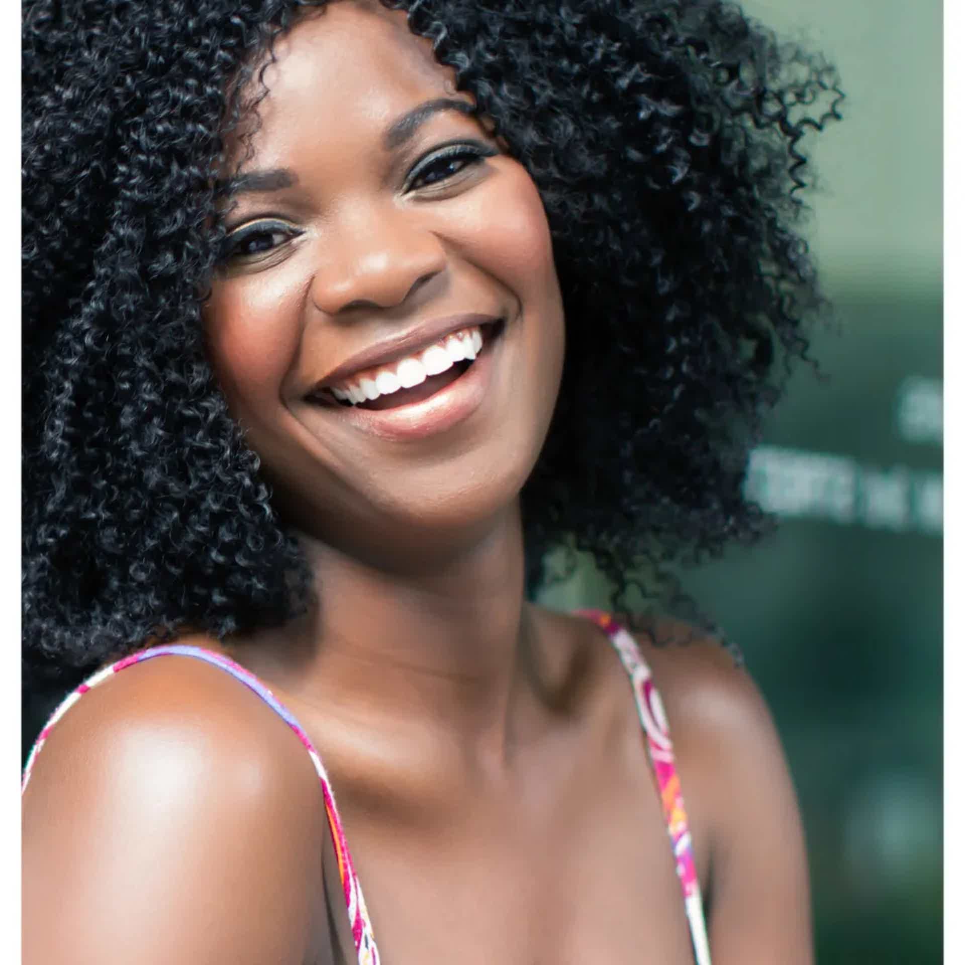 Smiling woman with curly hair wearing a colorful strap top, outdoor setting.