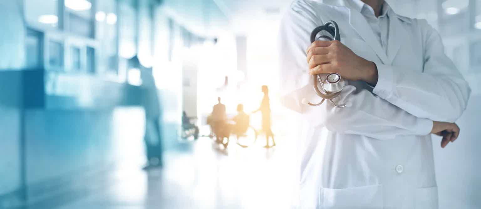 Healthcare worker in white coat holding stethoscope, standing in bright blue hospital hallway with blurred background.