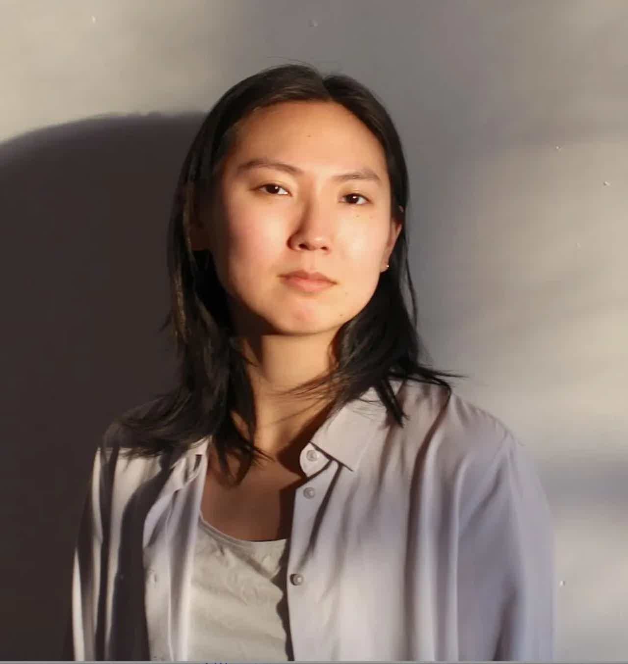 Portrait of a woman with shoulder-length dark hair, wearing a light shirt over a white top, neutral expression.