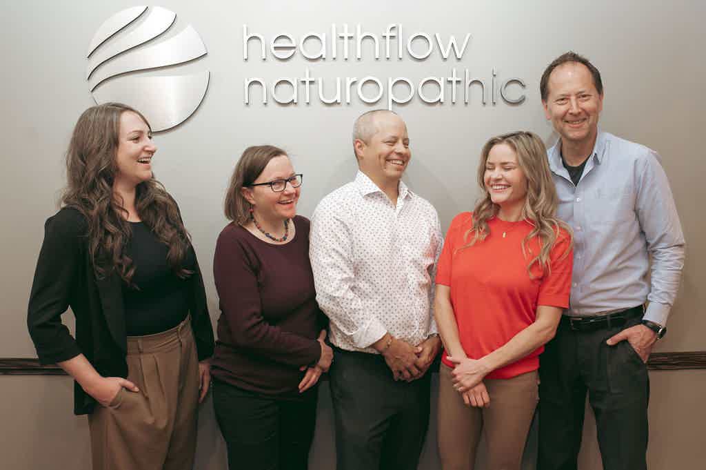 Smiling team standing in front of healthflow naturopathic logo wall, four women and one man posing.