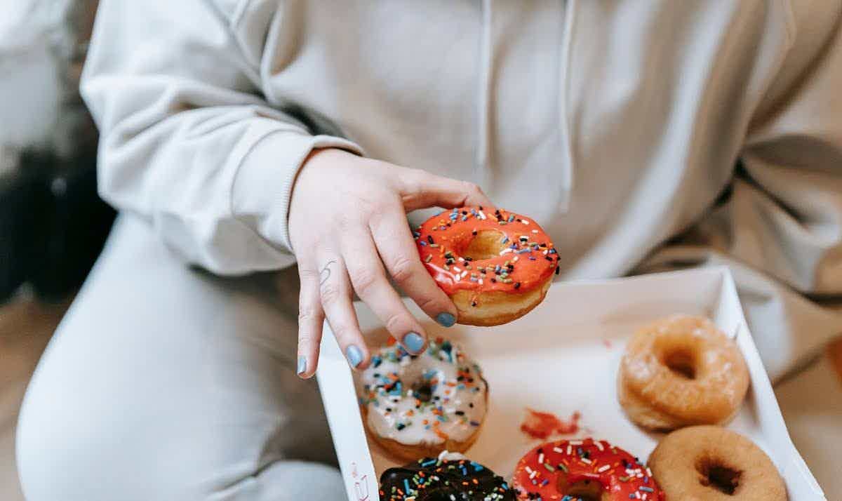 A person in a hoodie reaching for a brightly frosted donut from a box of assorted sprinkled donuts.