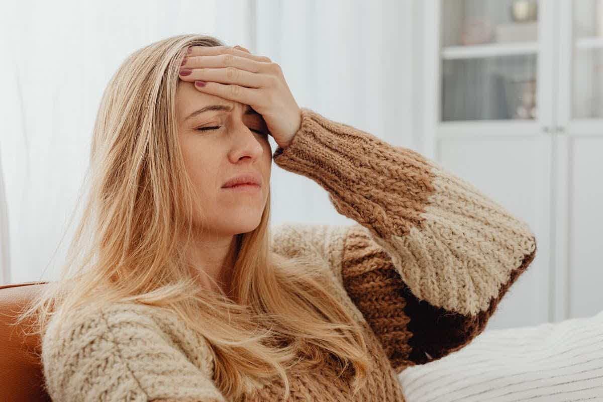 A woman in a knit sweater sitting on a couch with a hand to her forehead, looking exhausted and drained.