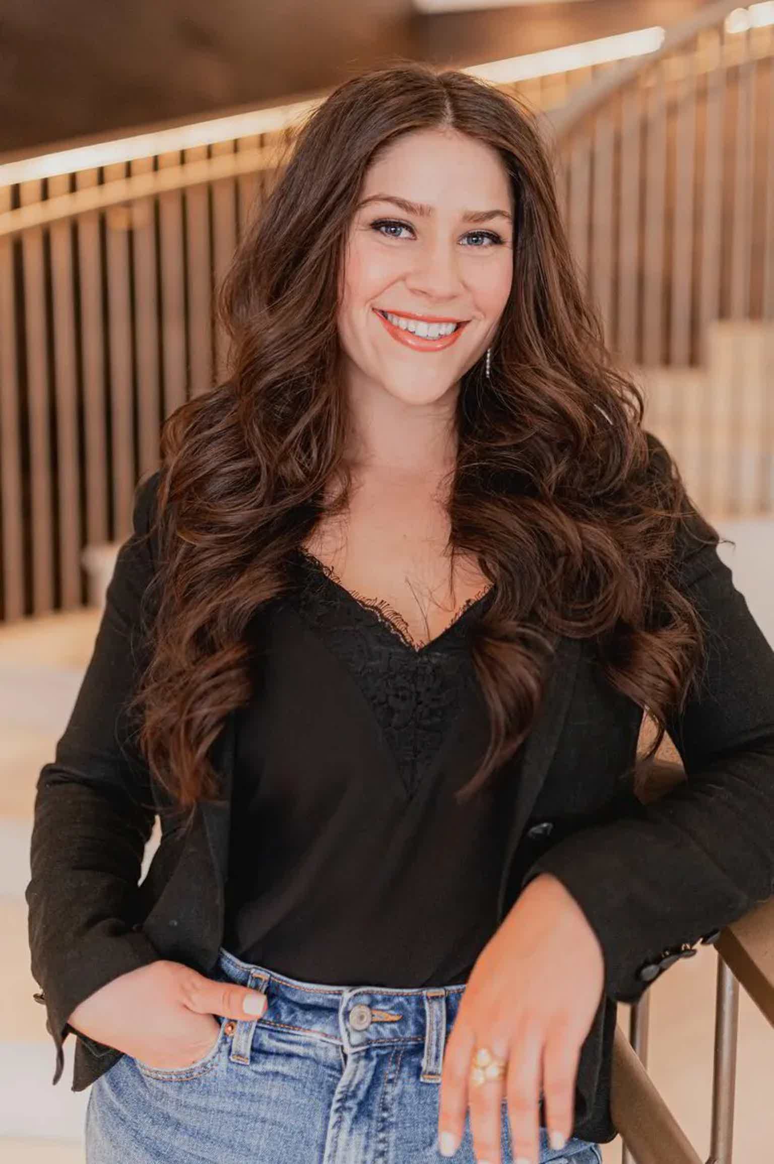 Smiling woman with long wavy brown hair leaning on a railing outdoors, wearing a black top and jeans.