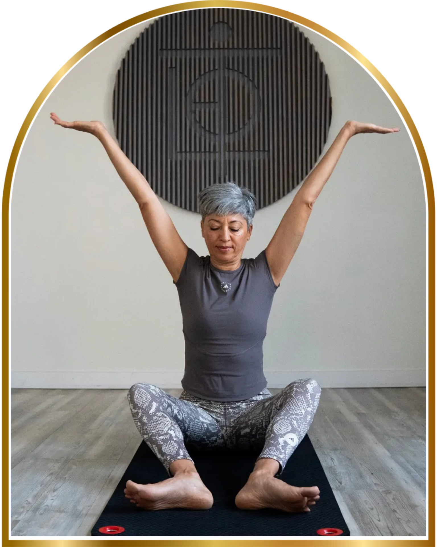 Person seated in yoga pose with legs crossed, arms raised, eyes closed, on mat against wall with dark backdrop.