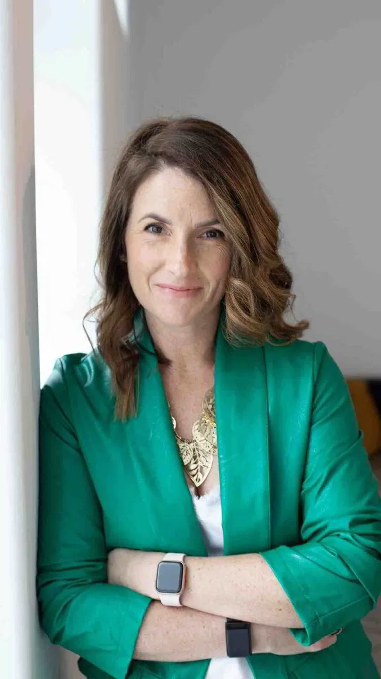 Woman in a green blazer with arms crossed, smiling at the camera indoors.