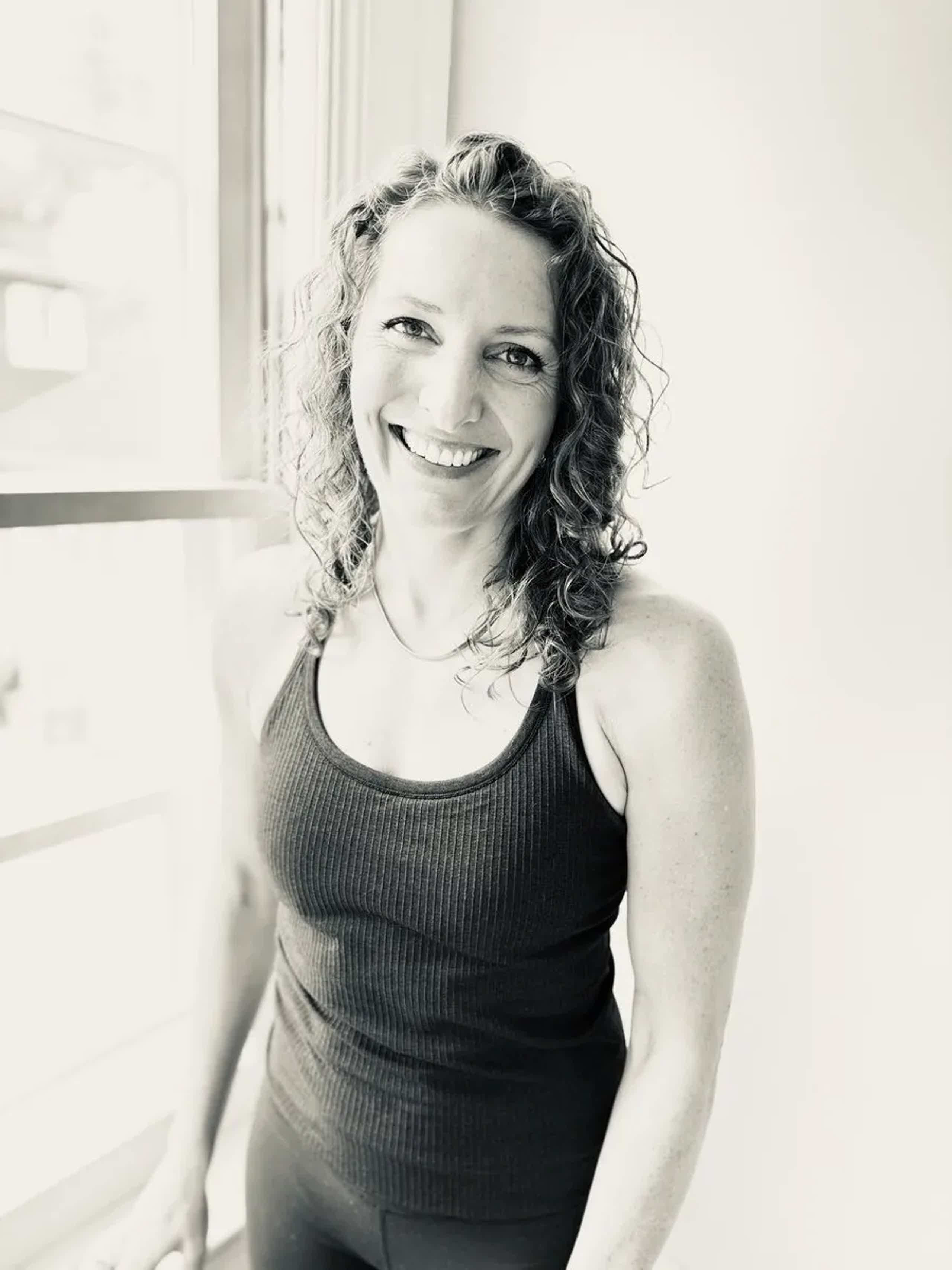 Smiling woman with curly hair wearing a black tank top, leaning by a window indoors.